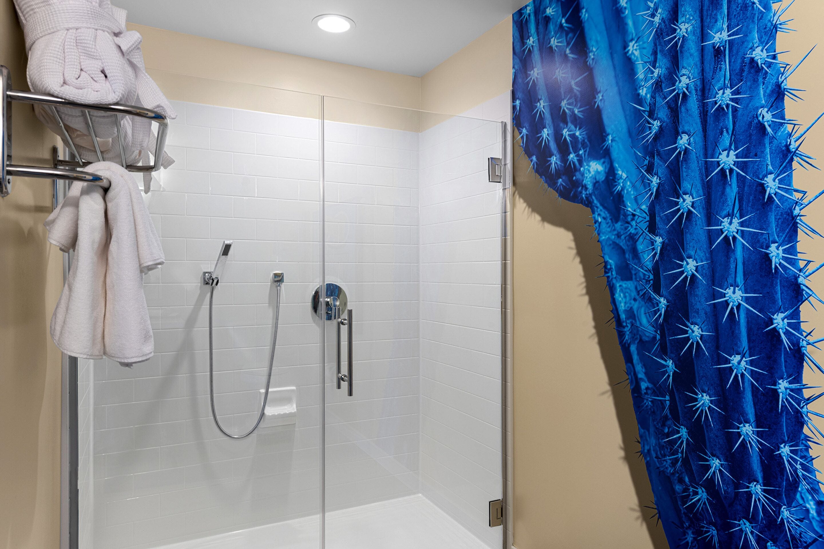 A shower with a glass door, a towel rack with folded towels, and a blue shower curtain with a cactus pattern.