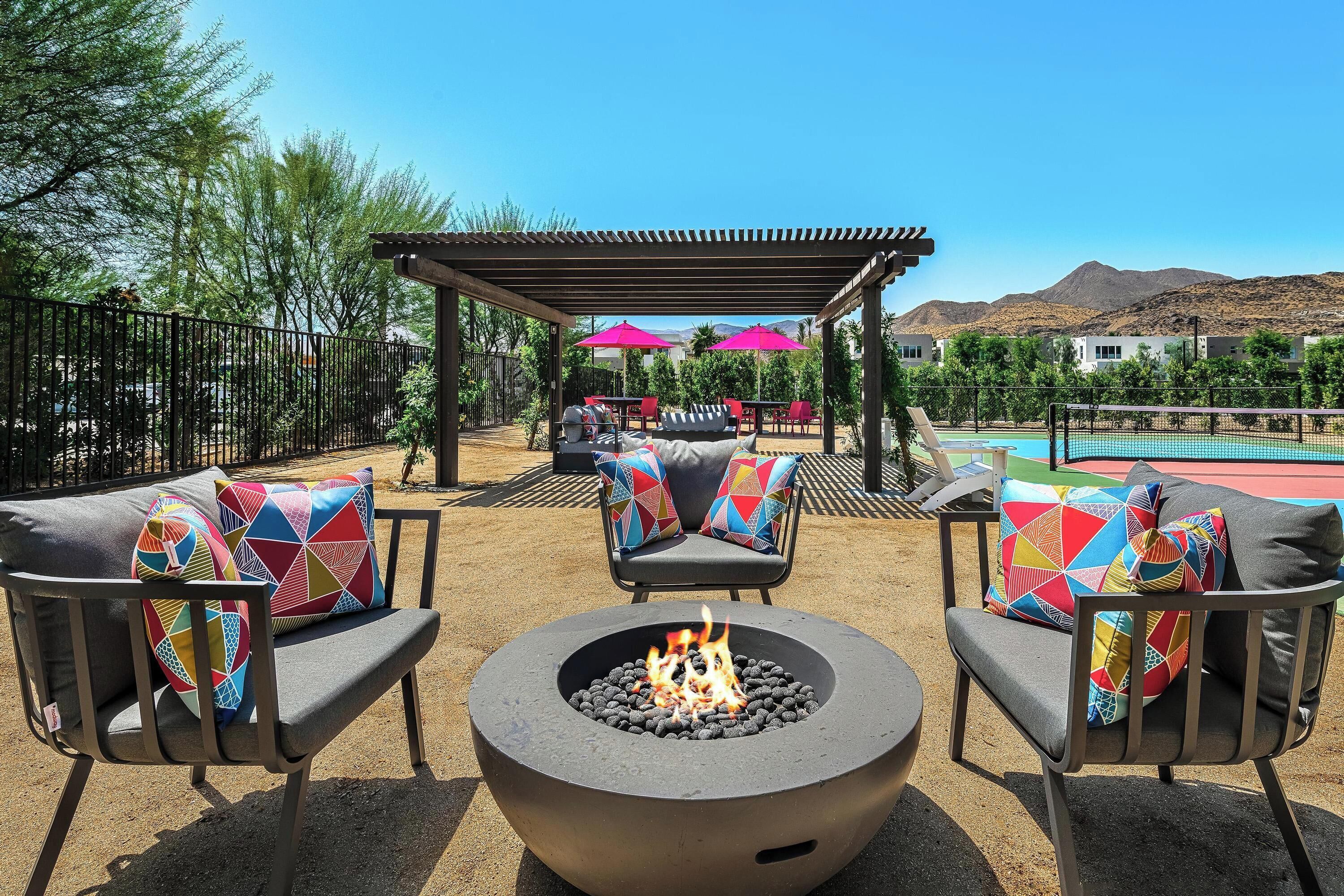 Un espacio patio con un brasero, dos sillas con cojines de colores, una pérgola con sombrillas y una piscina al fondo.