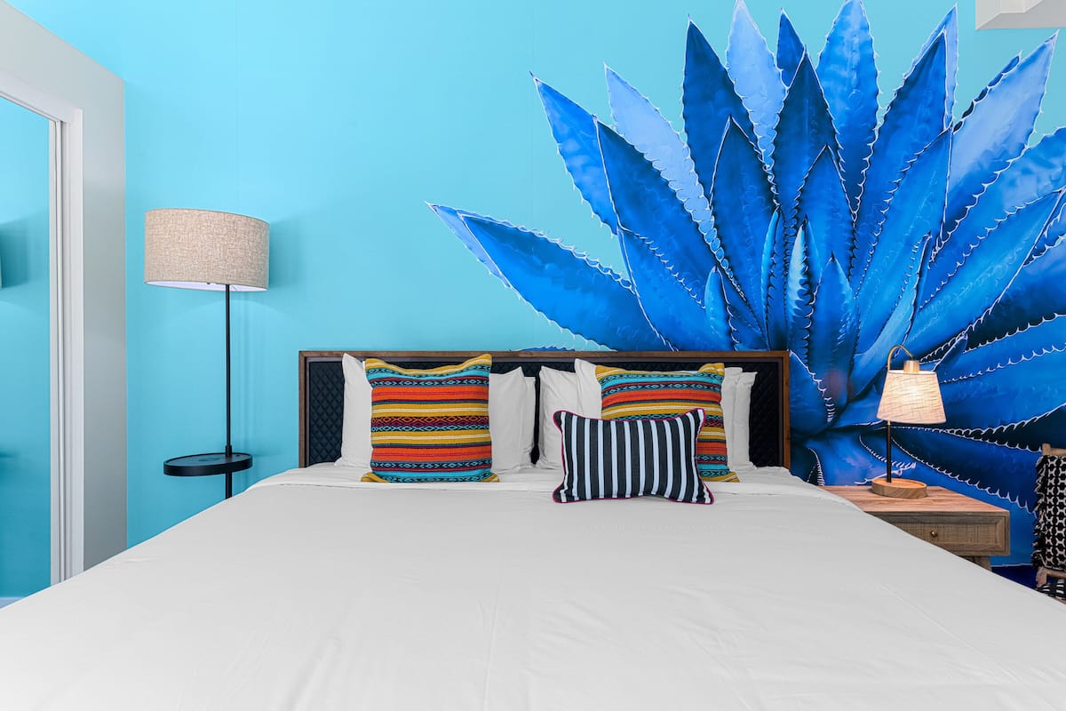 Een bed met wit beddengoed, kleurrijke gestreepte kussens en een grote blauwe plant op de achtergrond.