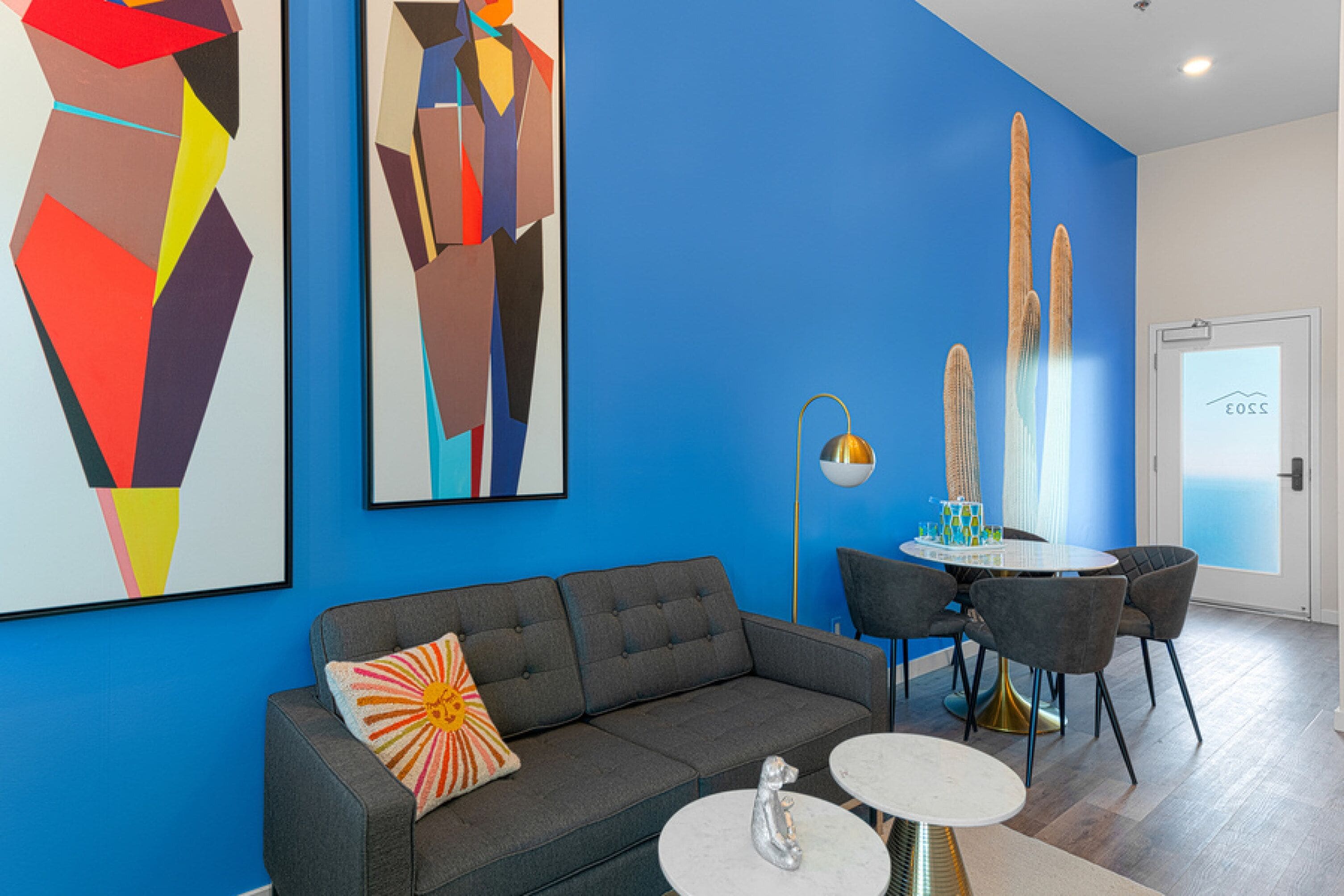 Un salon moderne avec un canapé gris, une petite table ronde et deux chaises. Des tableaux abstraits ornent le mur, et une porte dotée d’une poignée est visible.