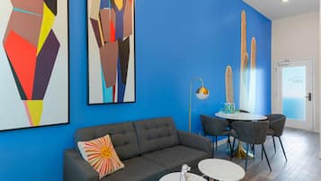Un salon moderne avec un canapé gris, une petite table ronde et deux chaises. Des tableaux abstraits ornent le mur, et une porte dotée d’une poignée est visible.