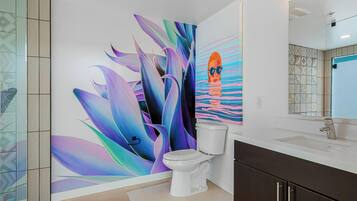 Une salle de bain au design moderne, avec des toilettes blanches, un grand miroir et un mur orné d’une fresque abstraite aux couleurs vives.