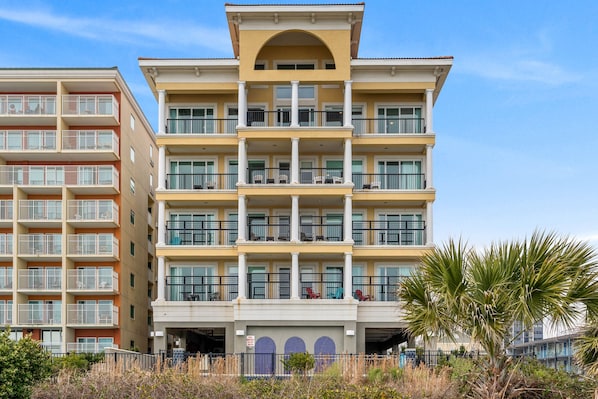 A multi-story hotel with balconies, a central entrance, and a parking area.