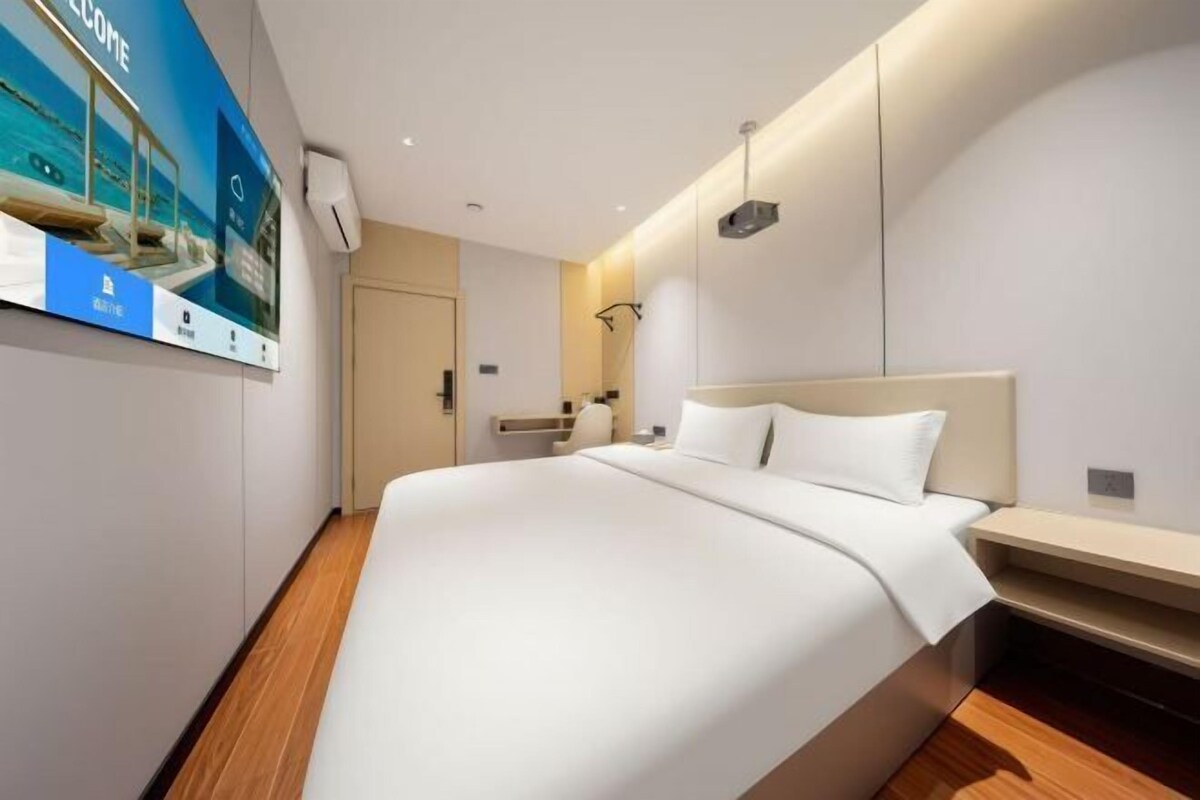 A modern hotel room with a large bed, a bedside table, a mirror, and a wall-mounted TV.