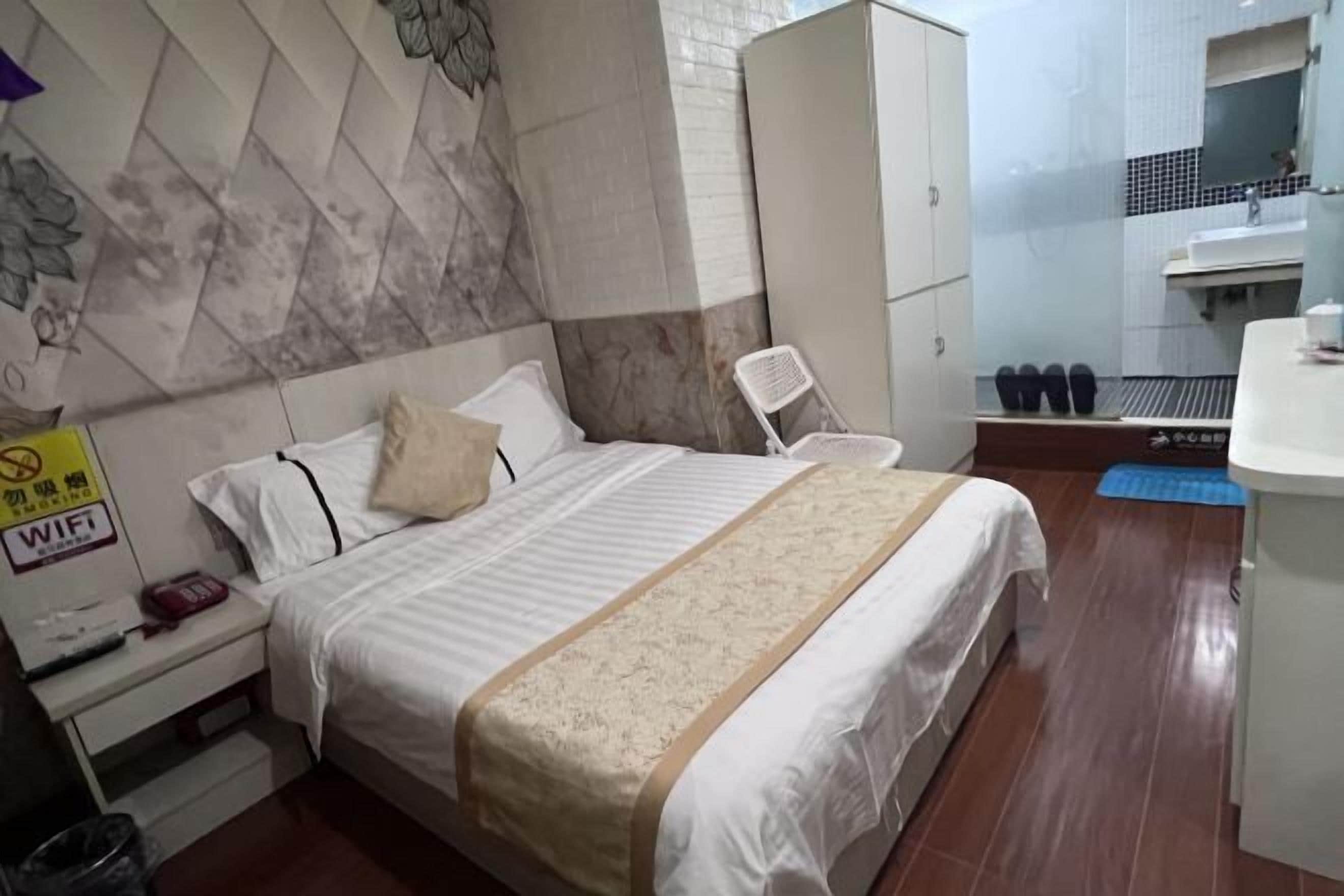 Une chambre d’hôtel comprenant un lit, une table de chevet, une armoire, une salle de bain avec un lavabo et un miroir, et un tapis bleu sur le sol.