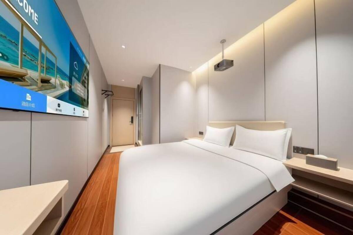 A modern hotel room with a large bed, a bedside table, a wall-mounted TV, and a wooden floor.