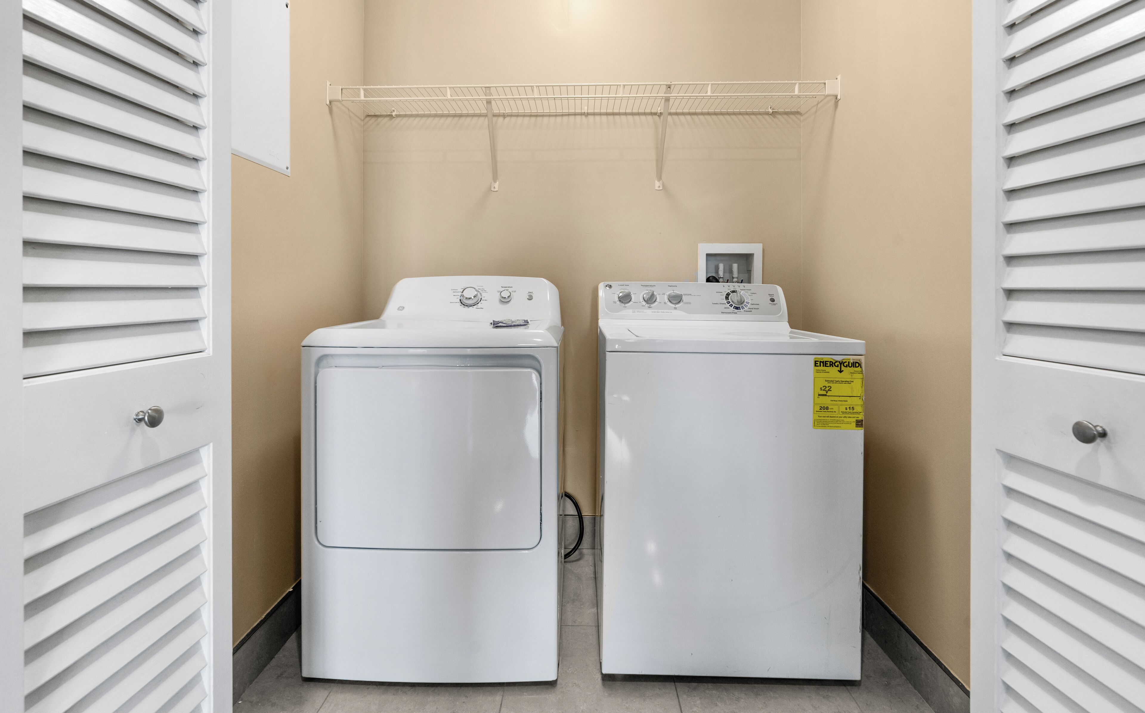A laundry room with two white washing machines and a dryer, a metal drying rack on the wall, and a door with a handle.