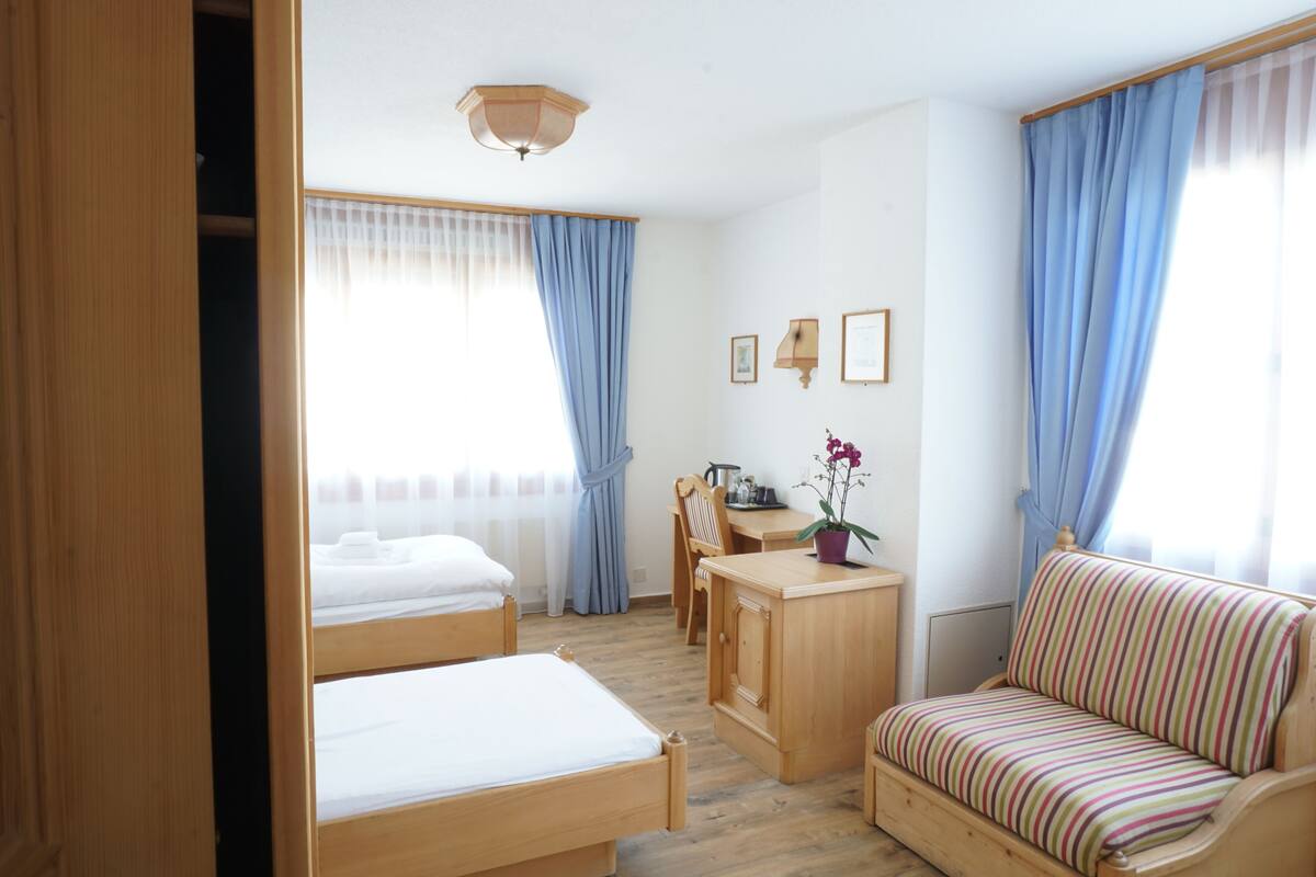 Habitación de hotel con dos camas de madera, un escritorio de madera, un sofá a rayas y una pequeña mesa con una planta en maceta.