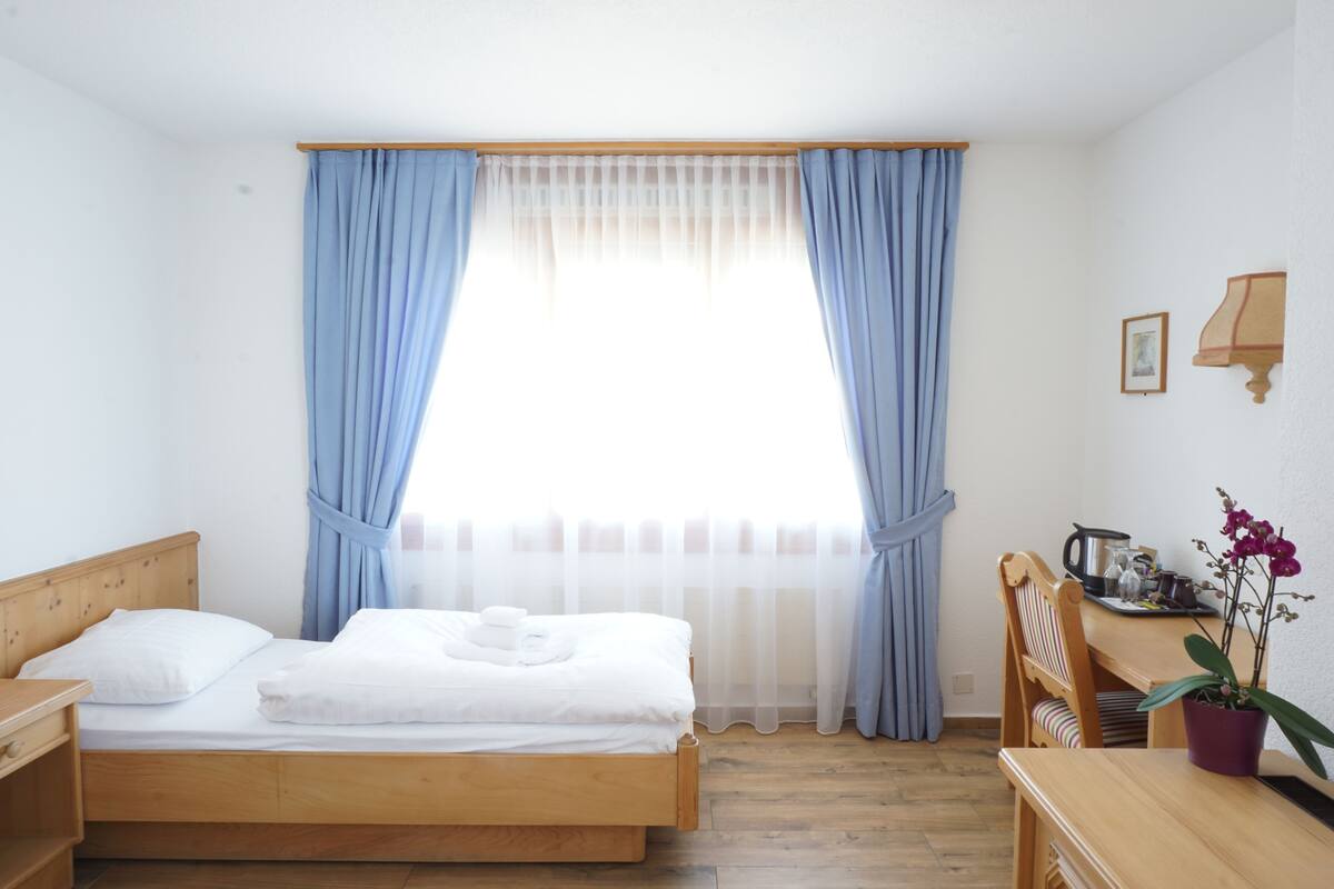 Un dormitorio con una cama de madera, un escritorio con silla, una orquídea en maceta y una ventana con cortinas azules.
