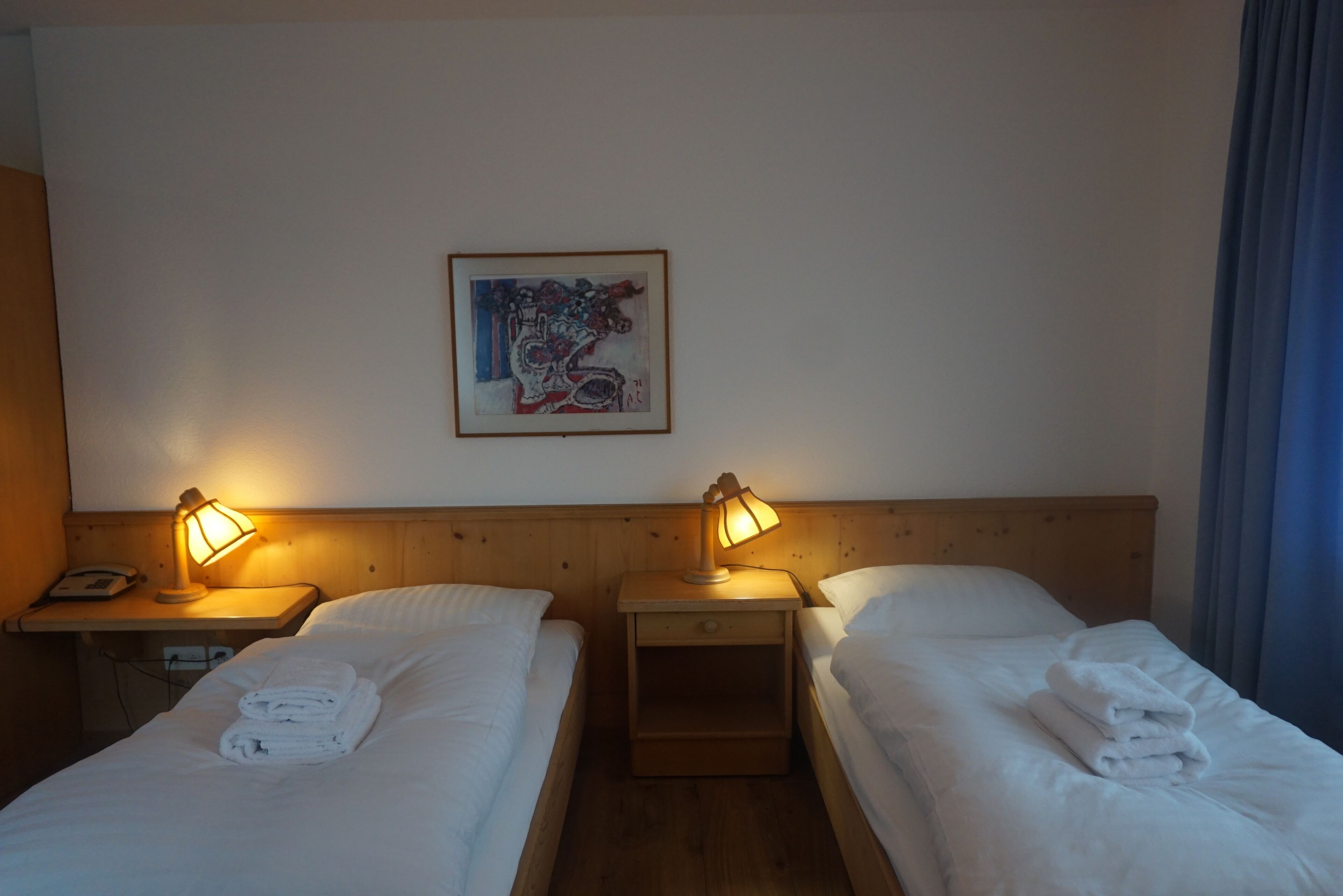A hotel room with two beds, each with a lamp and a folded towel. There is a painting on the wall and a wooden headboard.