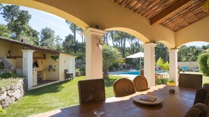Eine überdachte Terrasse mit Holztisch und Korbstühlen, mit Blick auf einen Swimmingpool und üppige Bepflanzung.