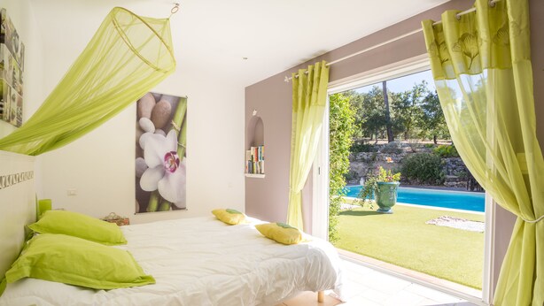 Ein Schlafzimmer mit Bett, grünen Kissen, einem Moskitonetz und Blick auf Pool und Garten.