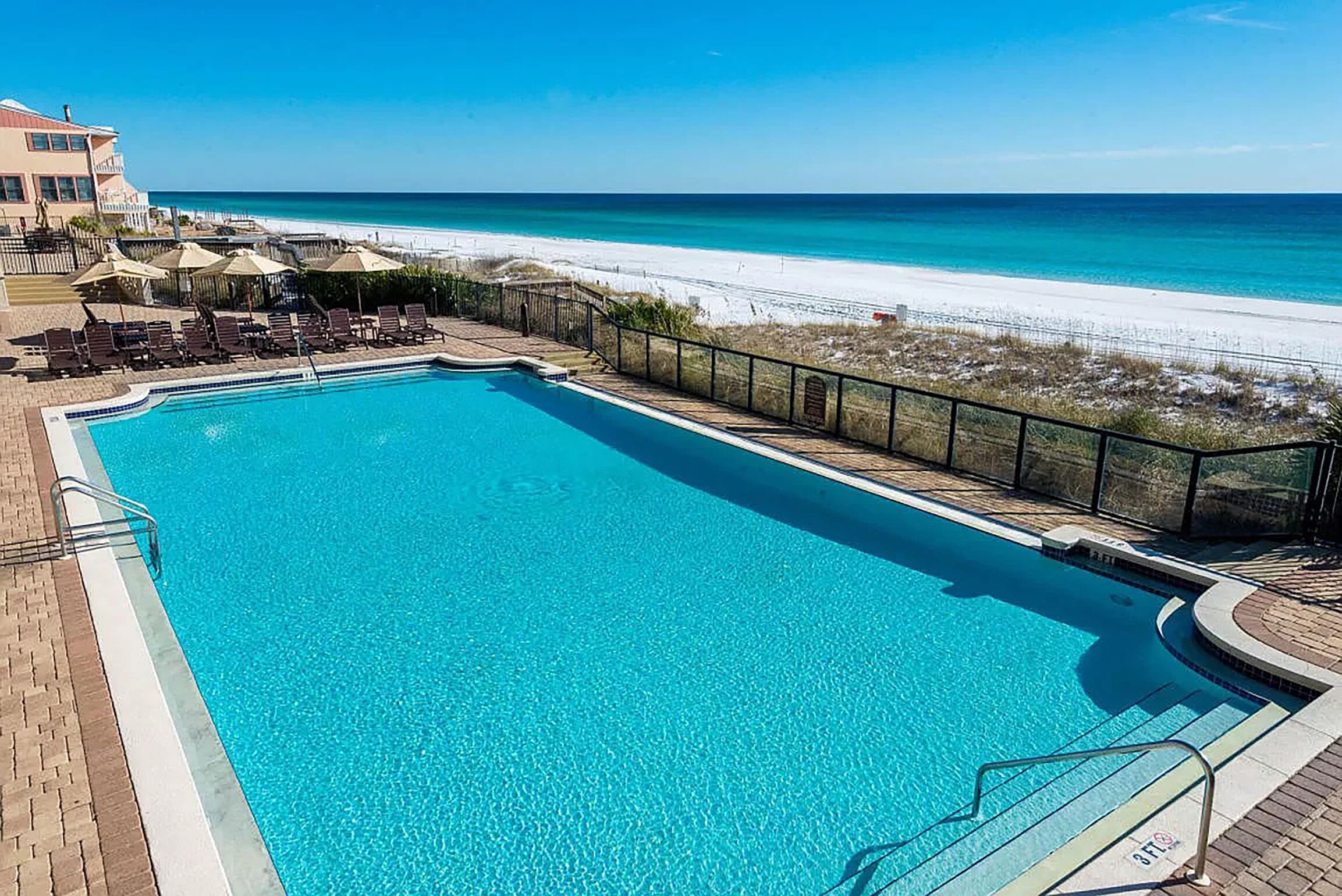 A clear blue pool with lounge chairs and umbrellas, a beach view, and a building in the background.