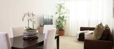 Een woonkamer met een bruine bank, een eettafel met stoelen, een televisie en een plant in pot.