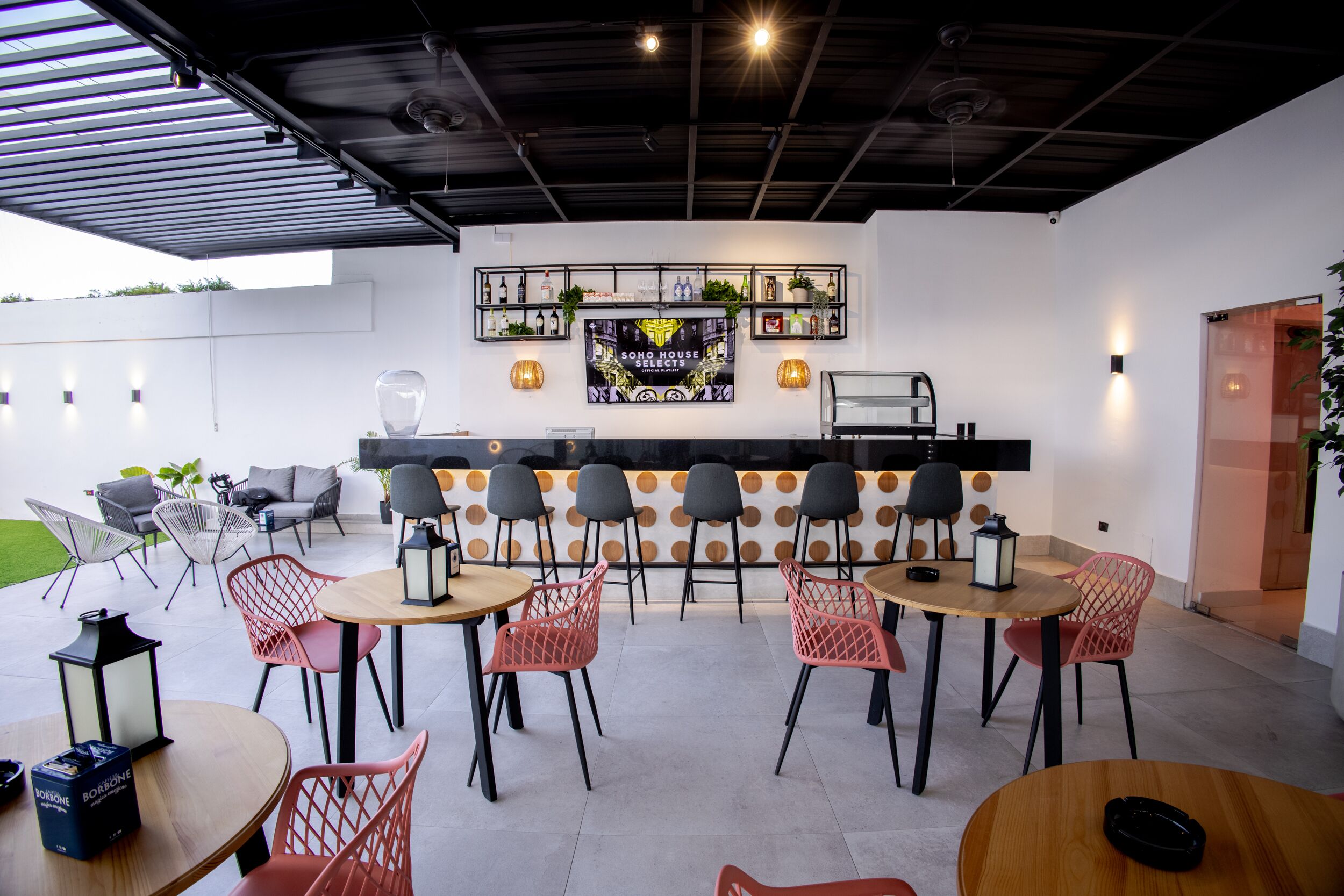 Un espace extérieur aménagé avec des tables et des chaises rondes, un bar proposant un choix de boissons et un panneau fixé au mur.