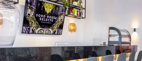 A bar area with a black countertop, four grey barstools, and a wall-mounted TV displaying 'SOHO HOUSE SELECTS'.