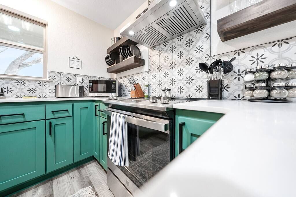 Een moderne keuken met turquoise kastjes, een magnetron, oven en een spoelbak. Boven de spoelbak is een raam en de achterwand is voorzien van een geometrisch patroon.