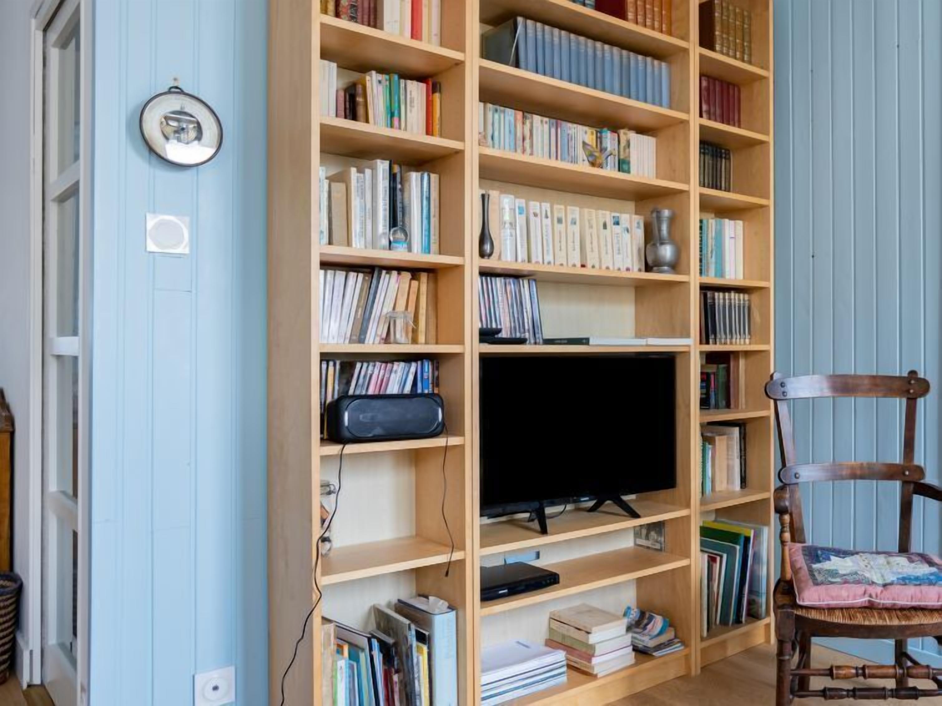 Une pièce comprenant une grande bibliothèque en bois remplie de livres et de DVD, un téléviseur posé sur un meuble, et une chaise en bois avec un coussin.