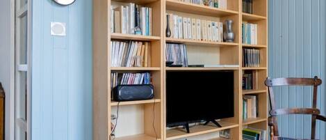 Une pièce comprenant une grande bibliothèque en bois remplie de livres et de DVD, un téléviseur posé sur un meuble, et une chaise en bois avec un coussin.
