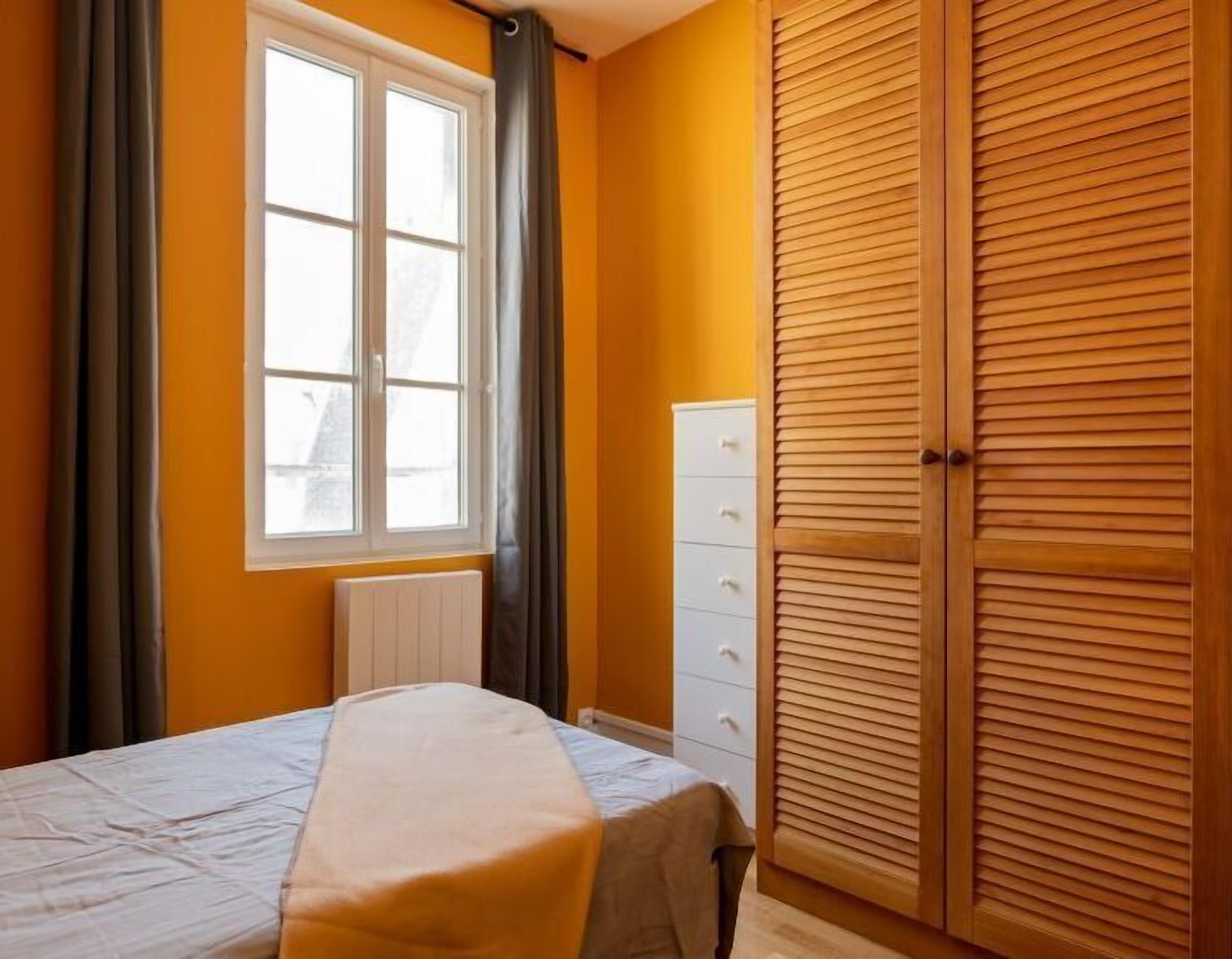 A bedroom with a bed, a radiator, a window with curtains, and a wooden wardrobe.