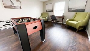 Una habitación con una mesa de fútbol de mesa, dos sillones verdes, una pequeña mesa con una silla y obras de arte en las paredes.