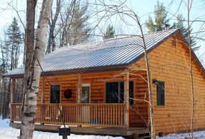 A wooden cabin with a porch, surrounded by trees and snow.