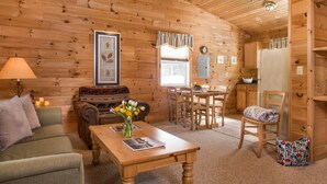 A cozy log cabin interior with a wooden dining table, a sofa, a coffee table with a vase of flowers, and a kitchen area with wooden cabinets. - Cozy Two Bedroom Cabin Near Smuggler's Notch (Jeffersonville)