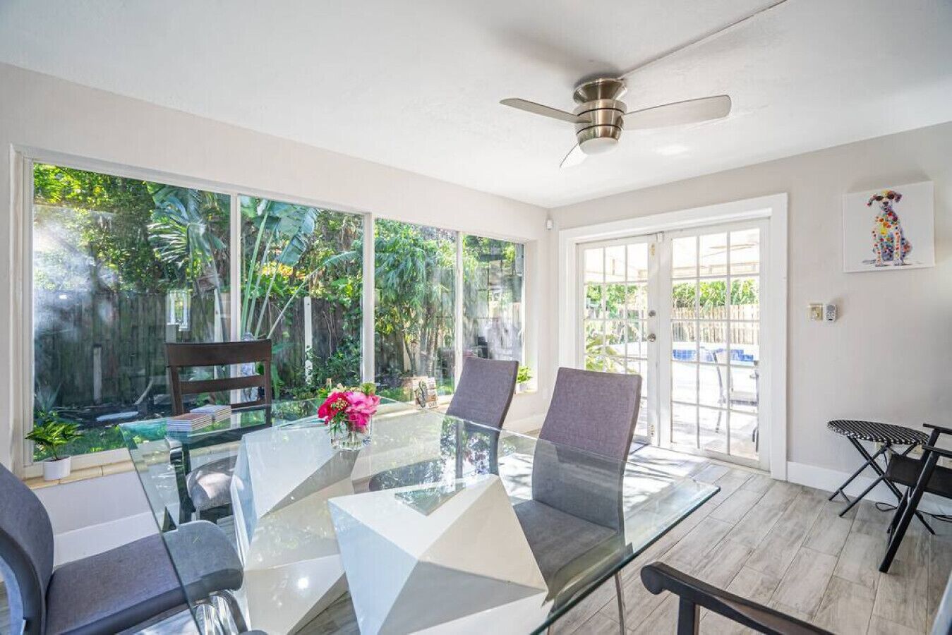A modern room with a glass table, two chairs, a ceiling fan, and a view of a lush garden through large windows.