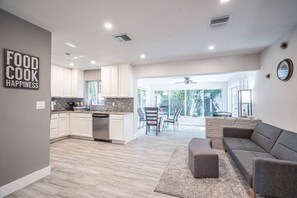 A modern kitchen with white cabinets and a gray countertop, a gray sofa, and a dining area with chairs. - Wilton Tropical Oasis with Heated Pool and Hot Tub (Oakland Park)