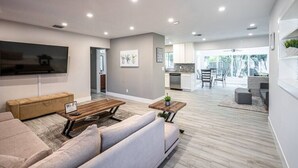 Een moderne woonkamer met een bank, salontafel en televisie. Deur open, zicht op een keuken met een eettafel en stoelen.