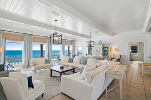 A spacious living room with white furniture, large windows overlooking the ocean, and a view of the sea.