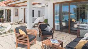 Un’area salotto esterna coperta con poltrone e divano, una piscina e vista sul mare.