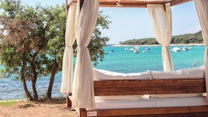 Cama de madera con dosel, número 005, con vista a una playa con barcos y agua azul cristalina.