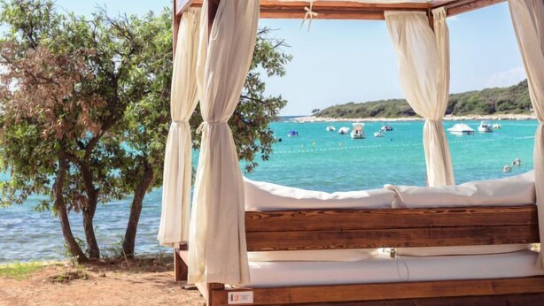 Ein hölzernes Bett mit Baldachin, Nummer 005, mit Blick auf einen Strand mit Booten und klarem, blauem Wasser.