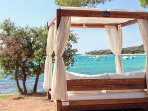A wooden bed with a canopy, number 005, overlooking a beach with boats and clear blue water. - Luxury Beachside Villa Porto Bus by Interhome (Rovinj/Bale)