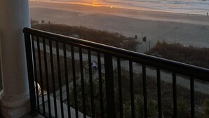 Vista del atardecer desde un balcón con barandilla, con vista a una playa y el océano.
