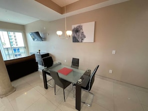 Uma sala de estar moderna com uma mesa de jantar de vidro, cadeiras pretas, um jogo americano vermelho e uma TV de tela plana na parede.