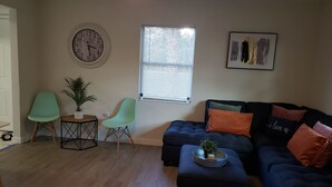 Un salon avec un canapé bleu foncé, deux fauteuils vert clair, une petite table sur laquelle est posée une plante, une horloge murale ronde, une fenêtre avec des stores et un tableau abstrait encadré.
