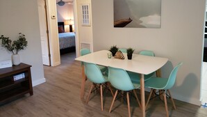 Un coin repas avec une table blanche et des chaises turquoise, un tableau au mur et une vue sur une chambre.