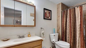 A bathroom with a wooden vanity, a white sink, a mirror with lights, a toilet, and a shower curtain. - Cypress Cottage in the heart of Steinhatchee! Fenced in yard! (Steinhatchee)