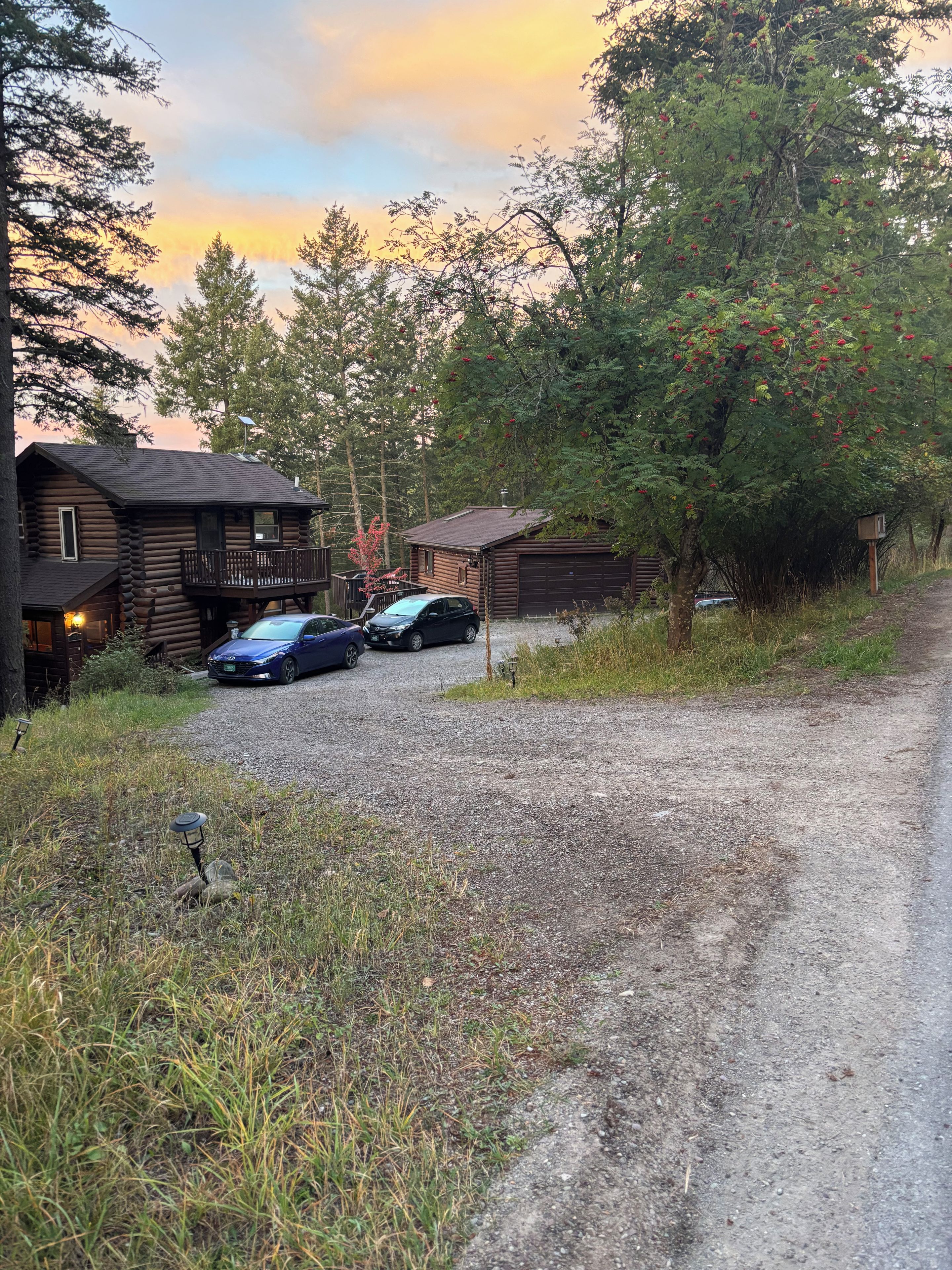 A rustic cabin with a gravel driveway, two parked cars, and a surrounding forest.