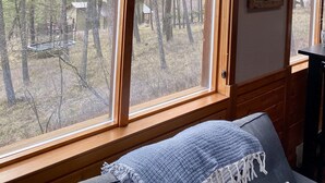 A room with a large window offering a view of a forested area, a bed with a blue blanket, and a wall-mounted picture.