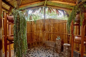 Una habitación circular con suelo de piedra, techo de bambú y paredes, con plantas colgantes y taburetes de madera rústica.