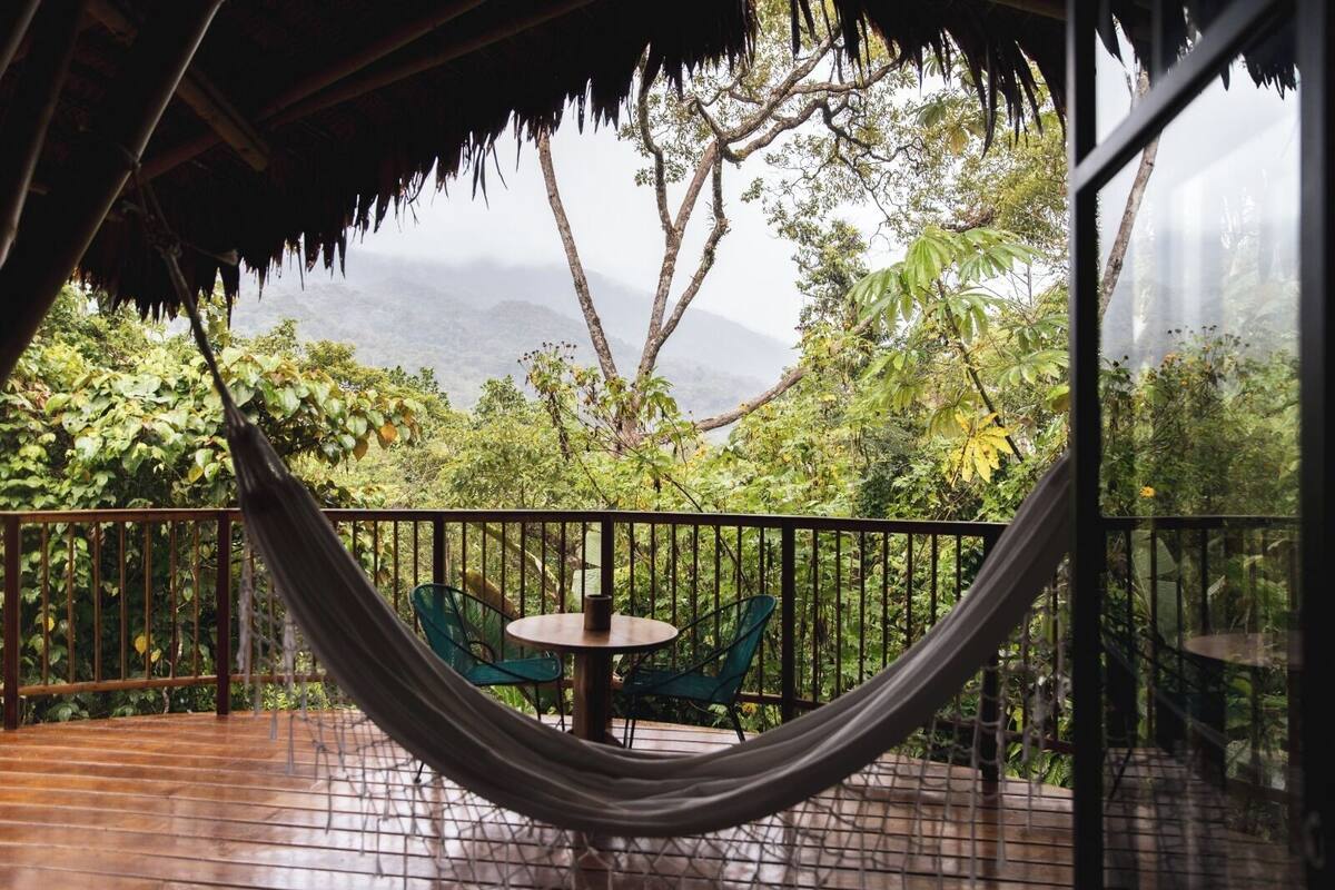 Een hangmat, een ronde tafel en stoelen op een balkon met uitzicht op een bebost berglandschap.