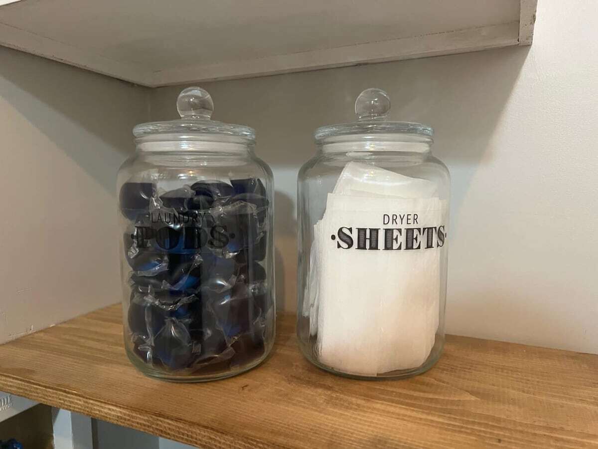 Dos frascos de vidrio en un estante, uno etiquetado 'Laundry Pots' y el otro 'Dryer Sheets'.