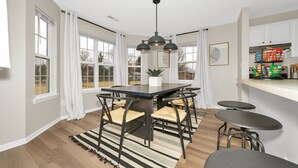 A modern kitchen with a dining area, featuring a large table, chairs, a hanging light fixture, and a kitchen counter with stools. - Luxurious Grand Park Family Home with Fenced Yard and Game Room! (Westfield)