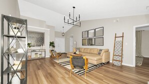 A spacious living room with a wooden floor, a beige sofa, a coffee table, and a chandelier. There is a bookshelf with decorative items and a ladder leaning against the wall. - Luxurious Grand Park Family Home with Fenced Yard and Game Room! (Westfield)