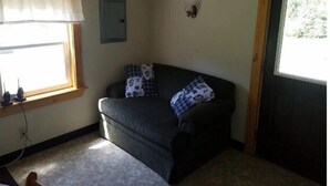 Una habitación pequeña, con poca luz, que presenta un sofá oscuro, una ventana con cortina, una lámpara de pared y una puerta.