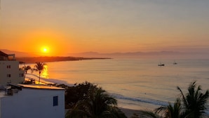 Une vue sur le coucher de soleil le long de la côte, avec une mer calme, des bateaux et un bâtiment doté d’un balcon surplombant l’eau.