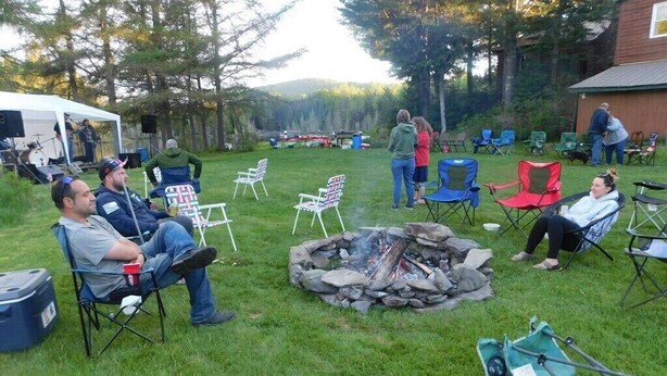 An outdoor gathering with people seated around a campfire, a tent, and various chairs in a grassy area. - 02) Northwoods Cabin (Stewartstown)